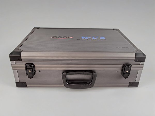 Closed electrical switch display case captured from the front, featuring a central black handle, two lockable latches, and a ribbed metal lid with printed branding.