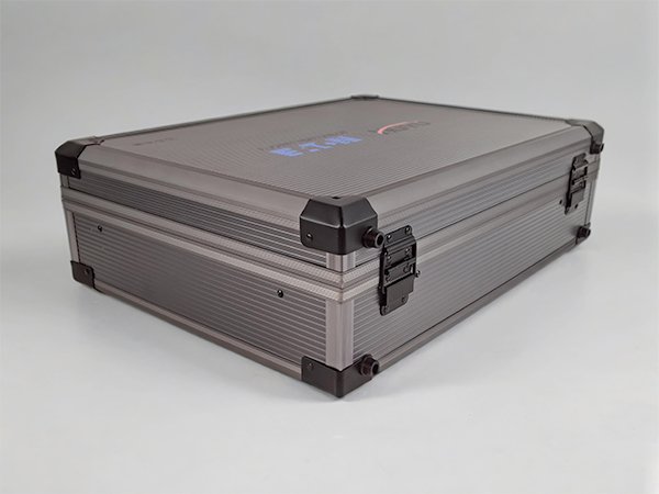 Closed gray aluminum electrical switch display case photographed from the rear corner, highlighting the ribbed metal surface, reinforced black plastic corners, and metal hinges.